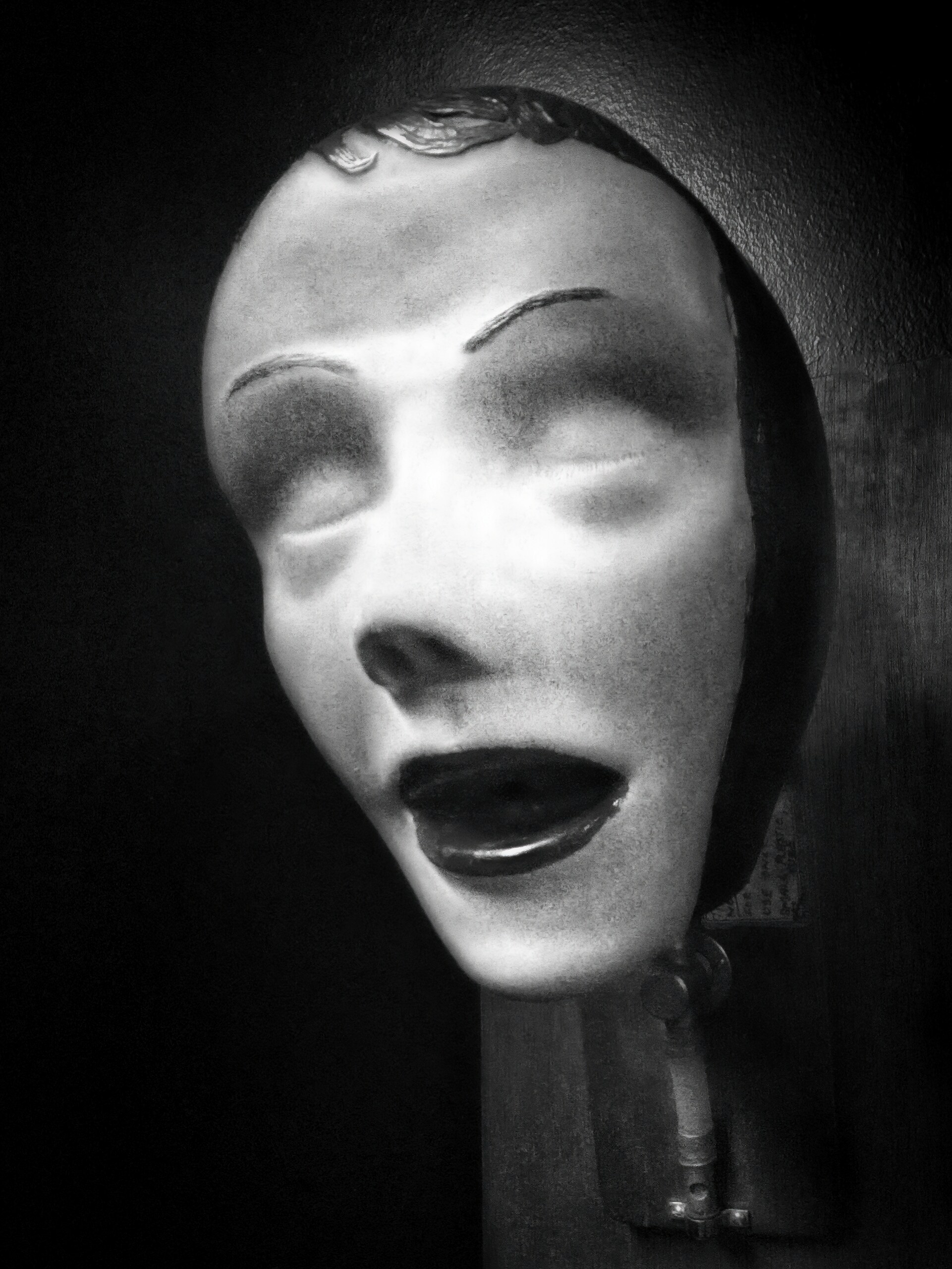 A pale, cracked mannequin face with an open mouth, emerging from shadow.