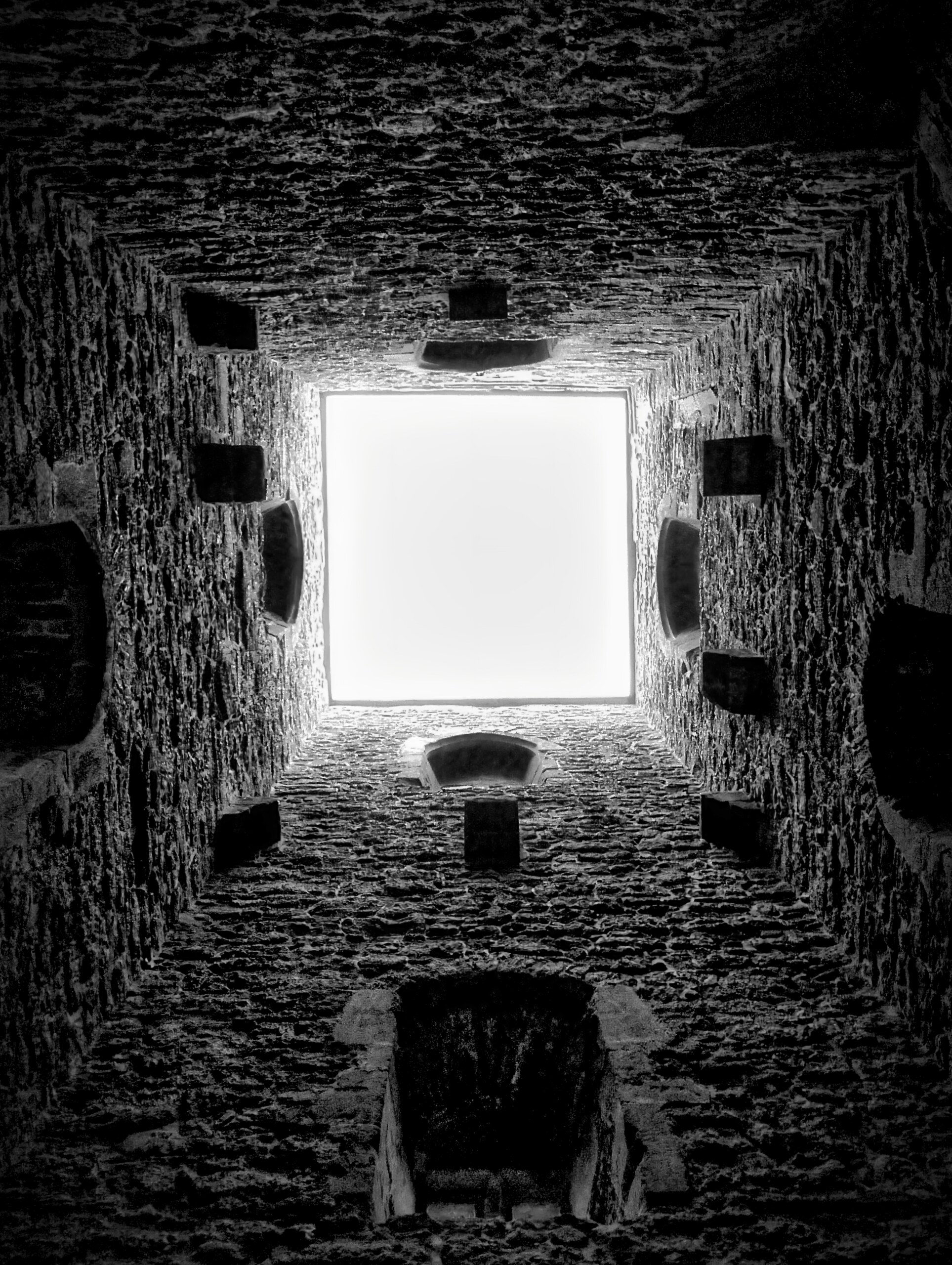Looking up through a square opening in stone toward a white sky.
